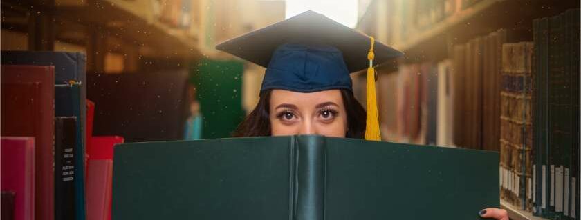 college student with graduation cap peeking over big book in library