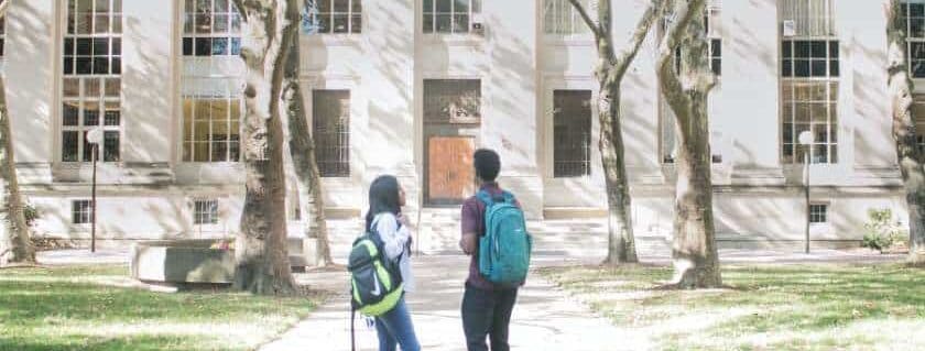 two people standing with their backpacks in front of a college building